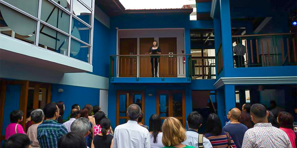 An actress standing behind the railing in an open-air corridor on the second floor of a blue building. A crowd of audience members looks up at her from the ground floor.