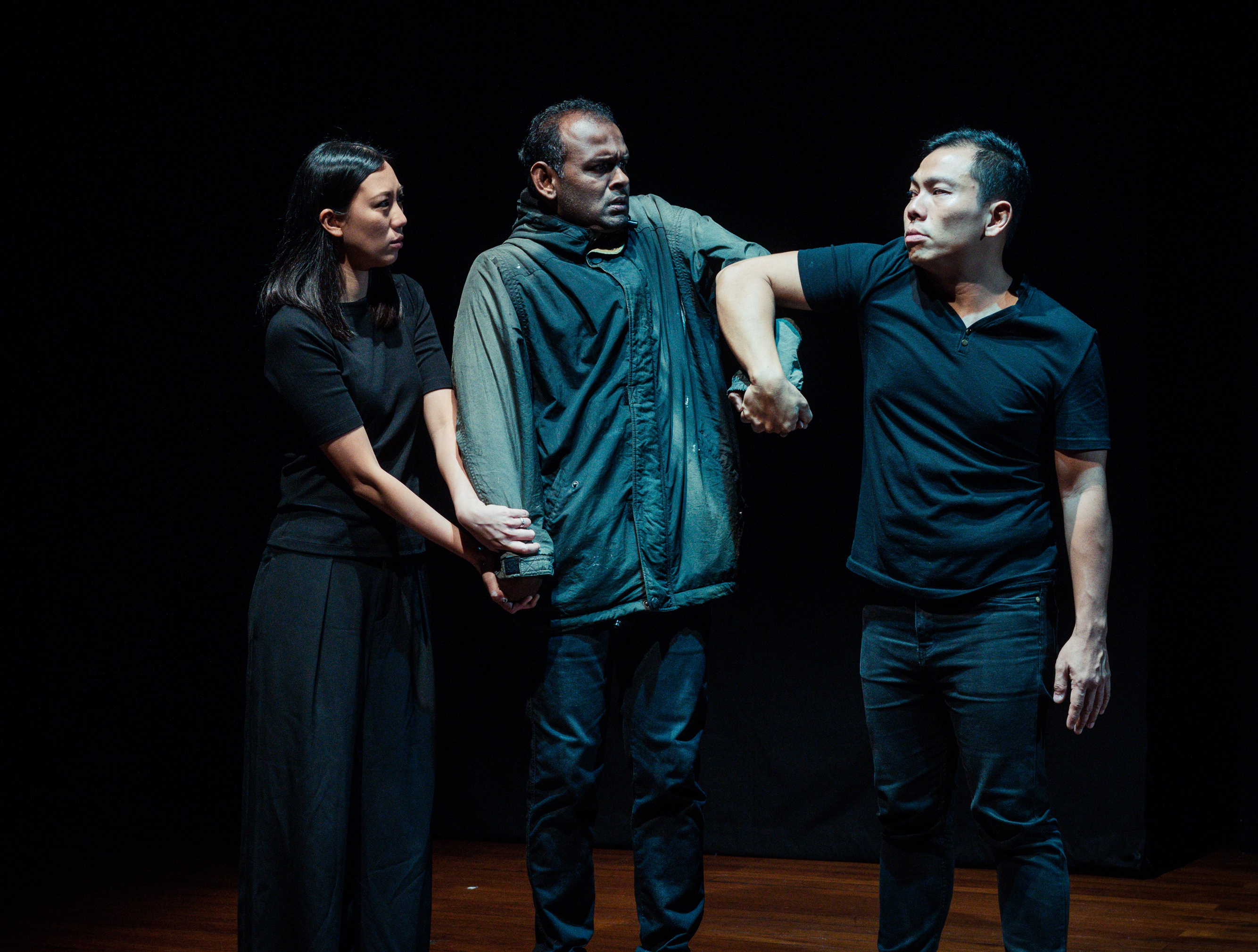 Two actors holding down the arms of a third actor standing in between them. He tries to pull away from them, looking upset.