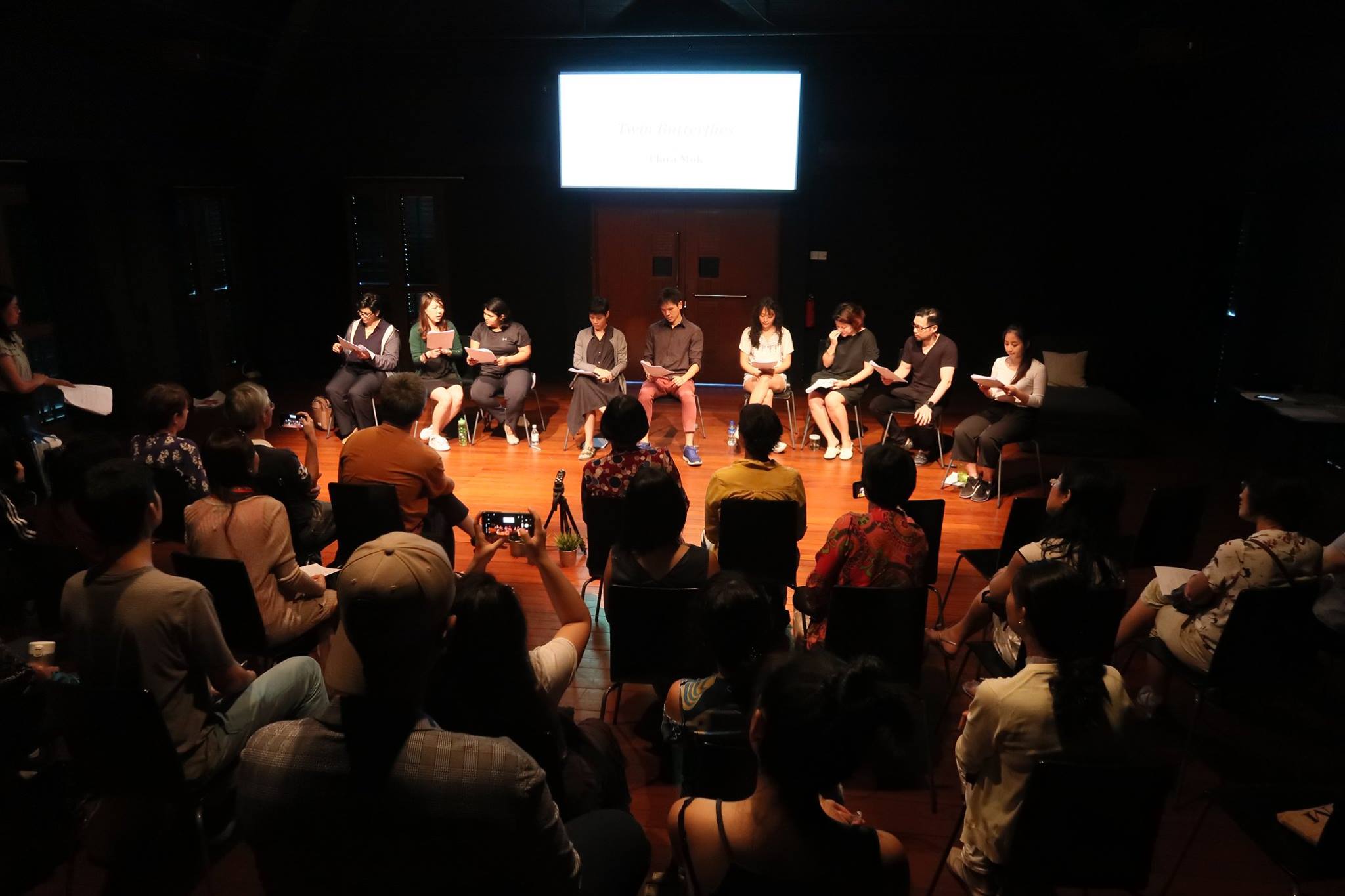 A row of performers reading a script in front of a large audience.