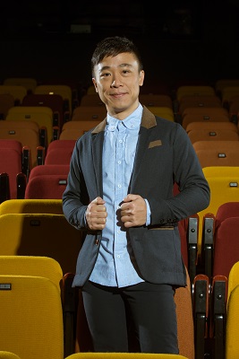 A male-presenting person in a blue shirt and grey jacket smiling into the camera. He is standing among red and yellow theatre seats.