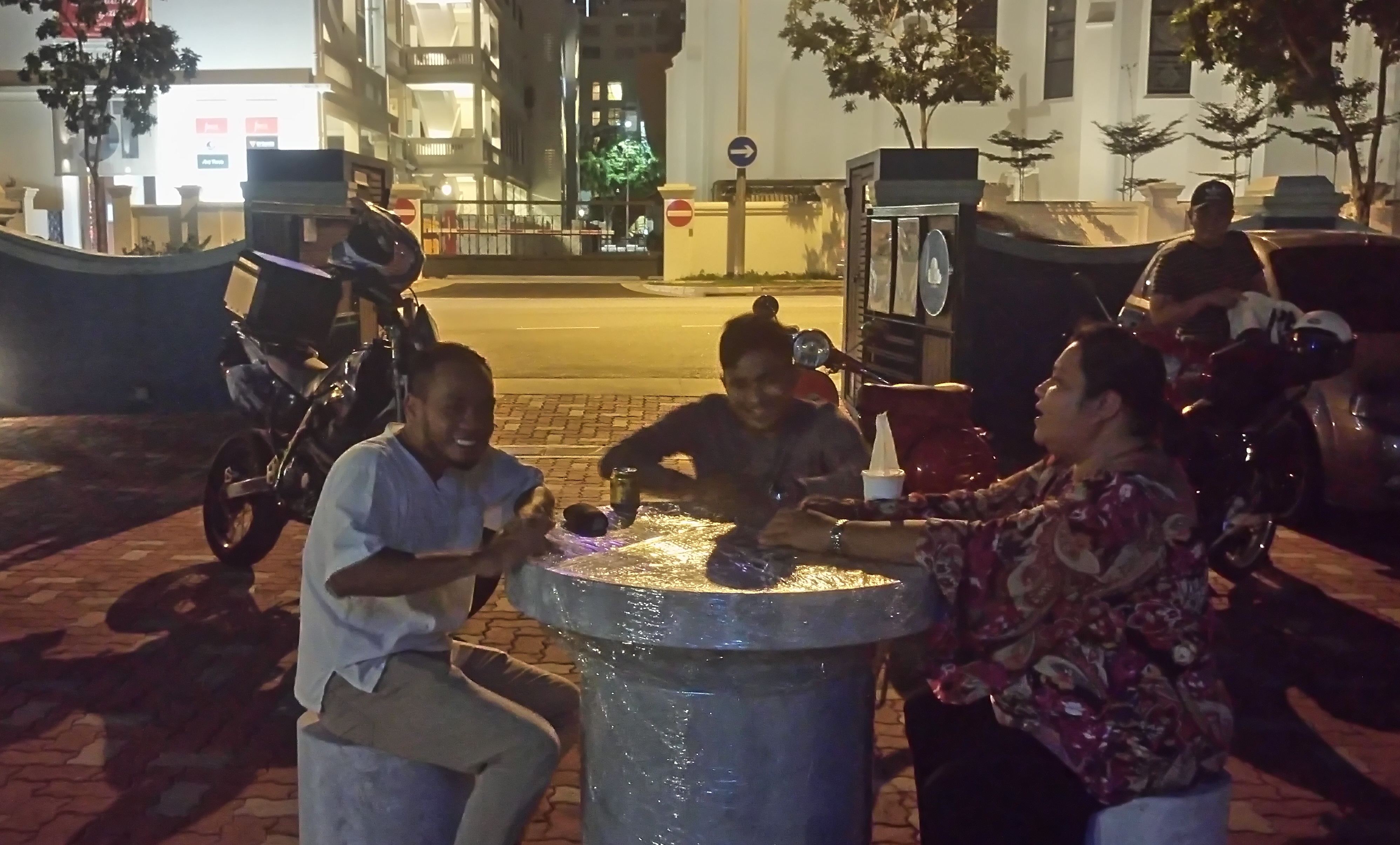A photograph of the actors of "Riders" in rehearsal. They are seated around a mock up of a stone park table at the courtyard of 42 Waterloo Street with their motorbikes in the background. 