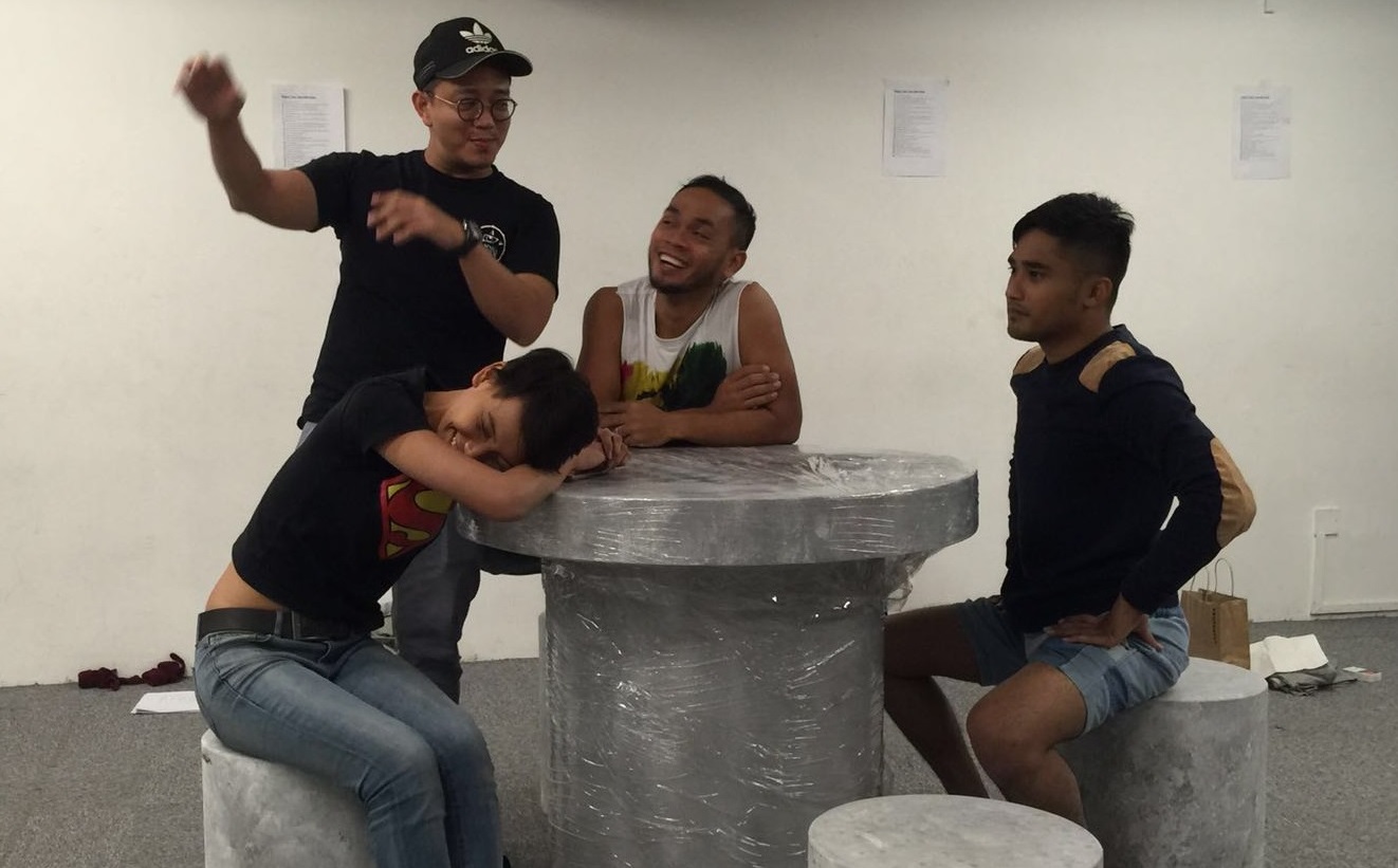 Production image of "Riders", with the four actors gathered around a public stone table and seats joking around.