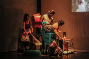 Three persons in a corner of a stage, surrounded by chairs and other household items. The person in the middle holds a large red plastic basin.