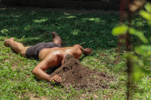 A person lies face-down on the grass with their head buried in a small mound of brown soil.