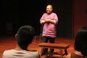 A male-presenting person in a pink shirt standing on a wooden stage, behind a low bench, smiling out an an audience. A male-presenting person in a pink shirt standing on a wooden stage, behind a low bench, smiling out an an audience.