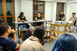A group of people seated in the 42 Waterloo Street Meeting Room, watching two performers at a table speaking to each other. A group of people seated in the 42 Waterloo Street Meeting Room, watching two performers at a table speaking to each other.