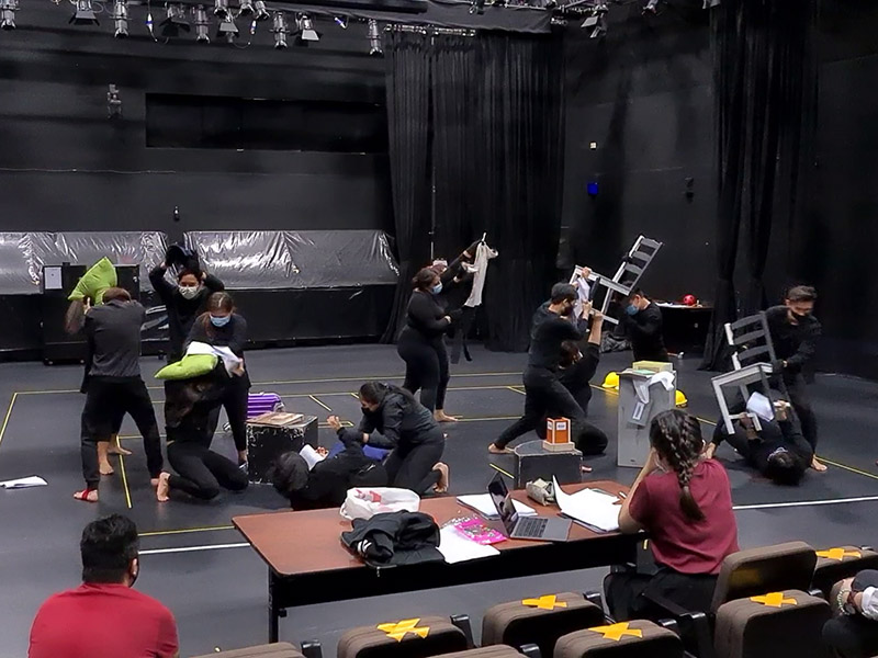 Several persons in black clothing in a black box, hitting each other with chairs and pillows. Watching them are two persons in red shirts, one of whom sits at a table with papers and a laptop in front of them.
