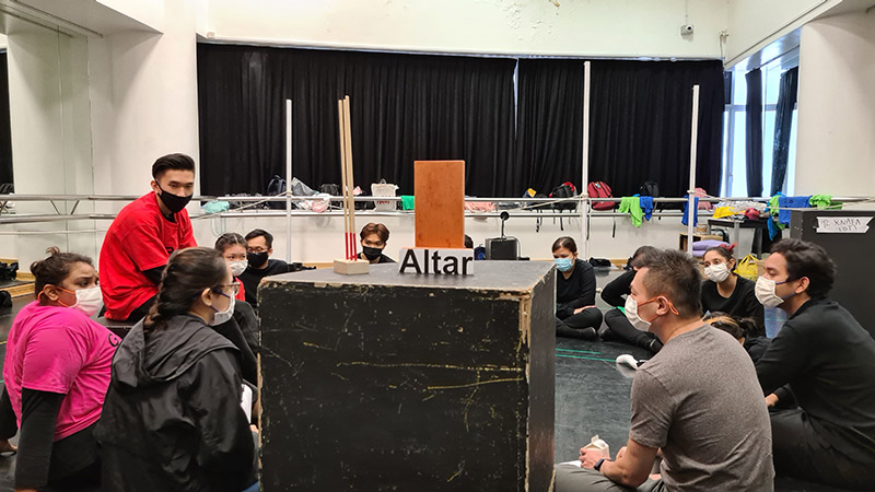 A crowd of people seated in a circle, speaking to one another. In front of them is a tall black box with a sign on top that reads 'Altar'.