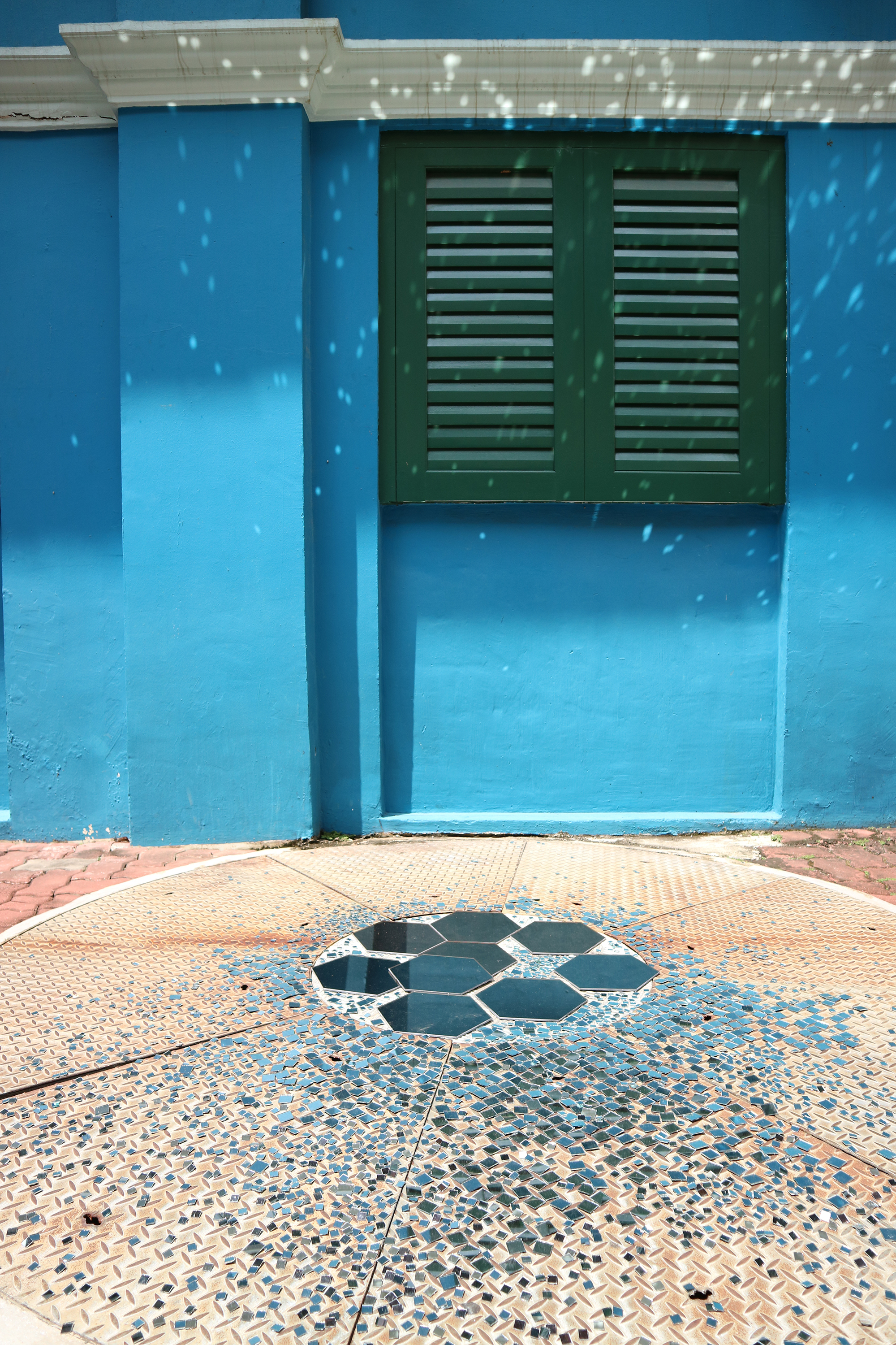 Small blue tiles scattered across a manhole cover along with larger hexagonal tiles. The tiles reflect light off them onto the facade of the Centre 42 building, creating a kaleidoscopic effect.