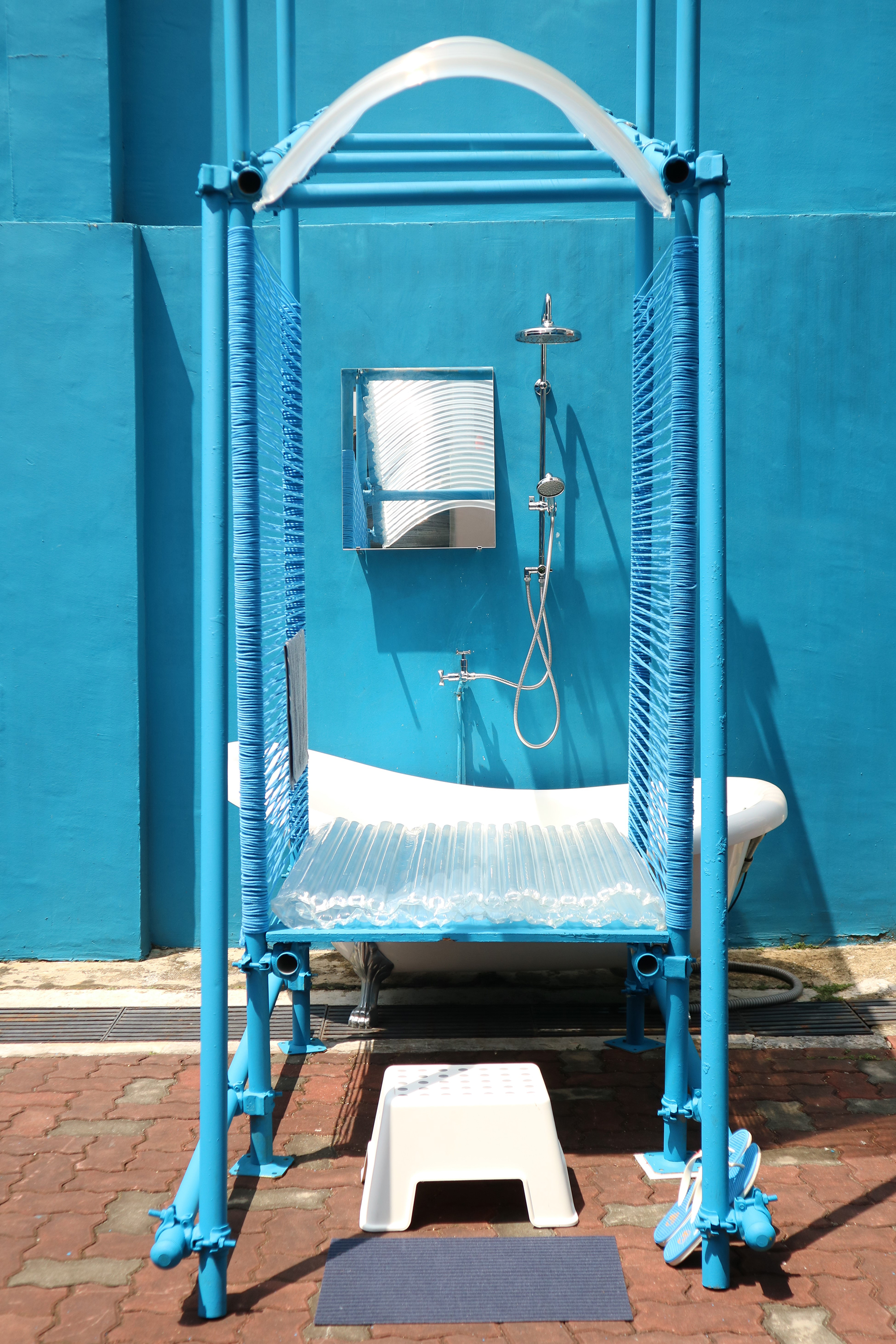 An installation of a shelter structure made out of pipes. A bath mat and small white stool line the entrance, with a pair of flip flops to the right. This leads to a white bath tub and shower head.