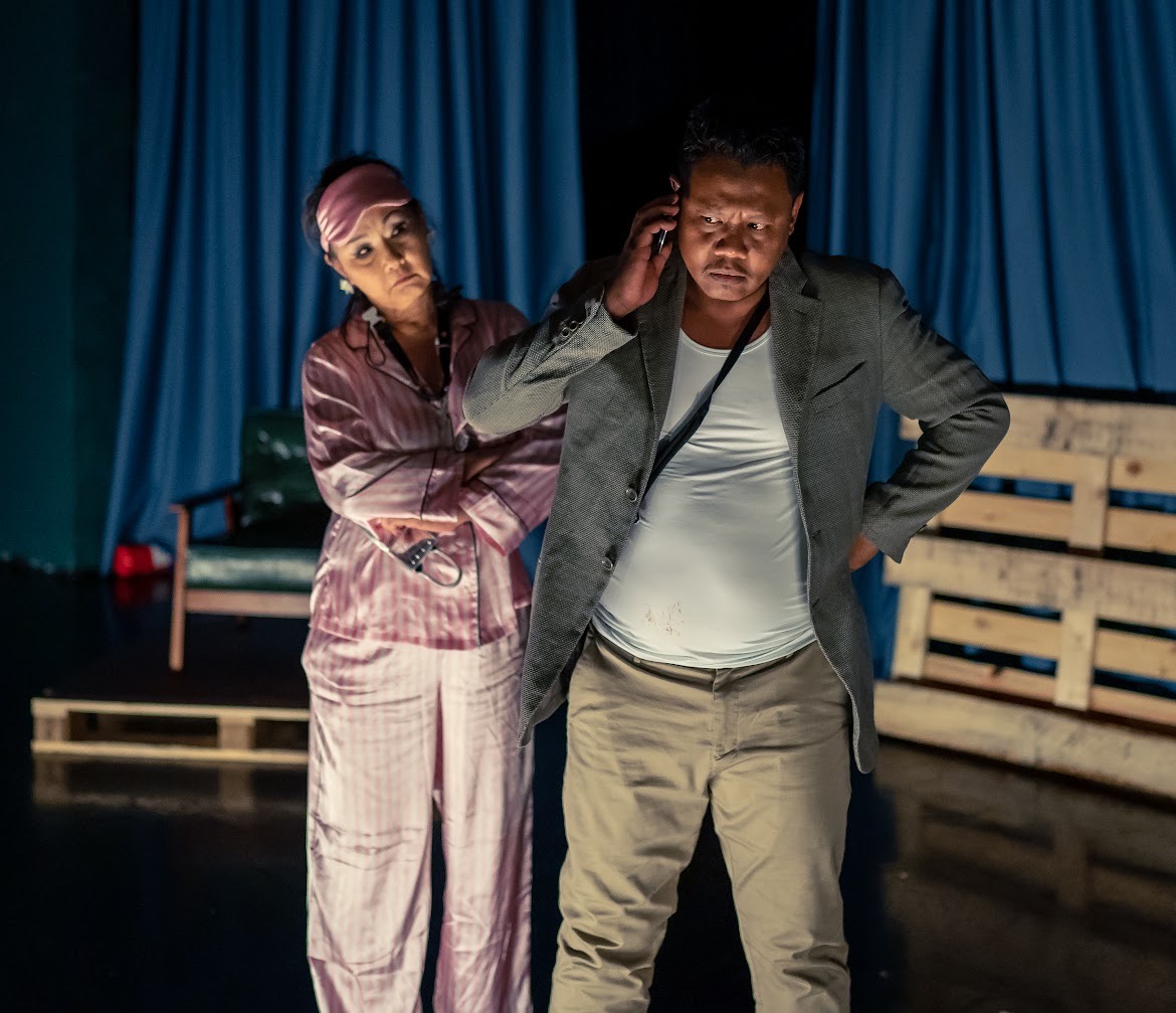 A male-presenting person is holding a phone to his ear with a female-presenting person with her arms crossed behind him.