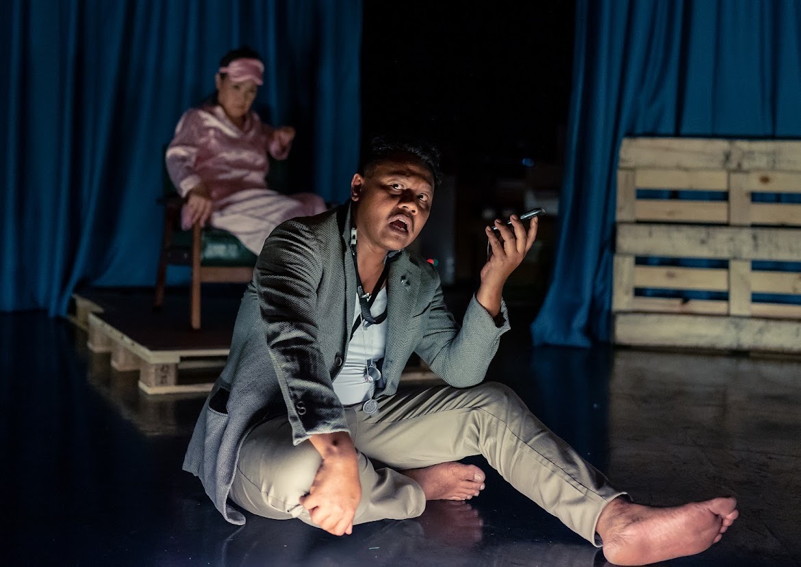 A male-presenting person is speaking into a phone in his hand with a female-presenting person staring at him on a sofa chair behind him.