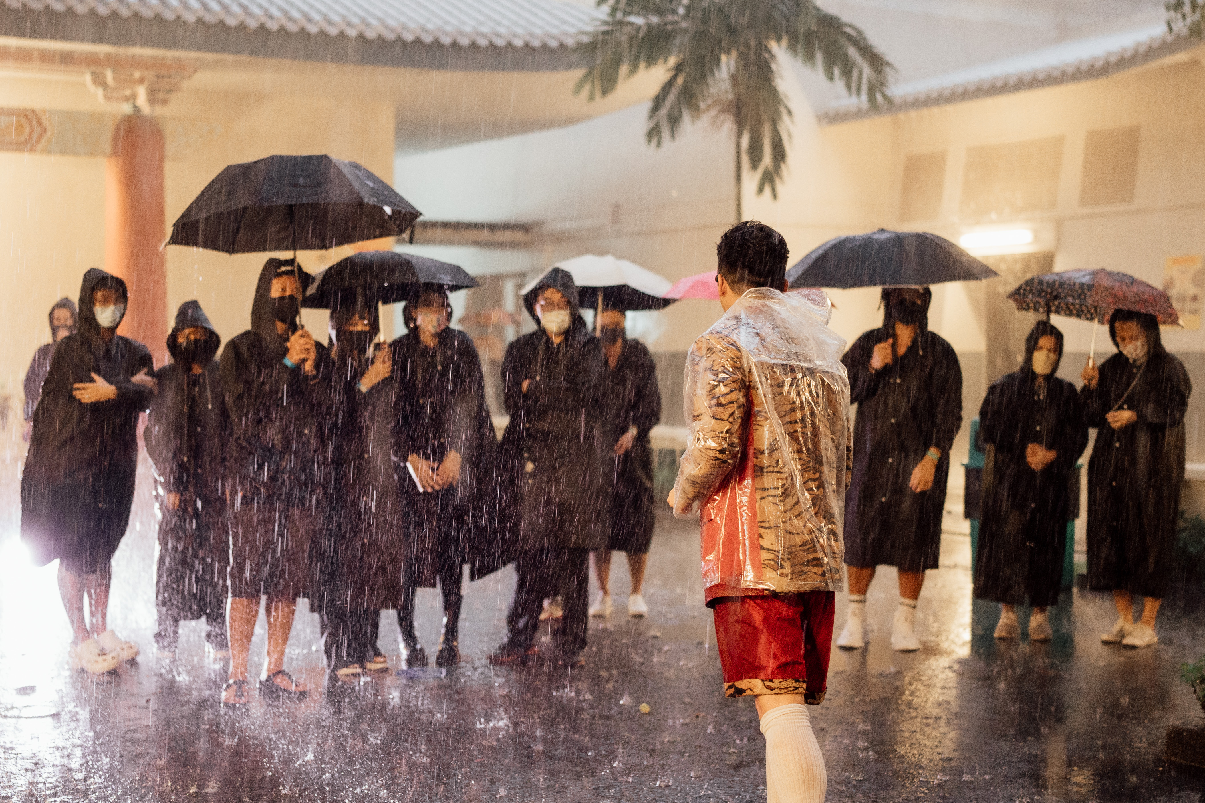 A group of people in black raincoats, standing under umbrellas in the rain. Facing them is a person in a shirt with a tiger print, speaking to them.