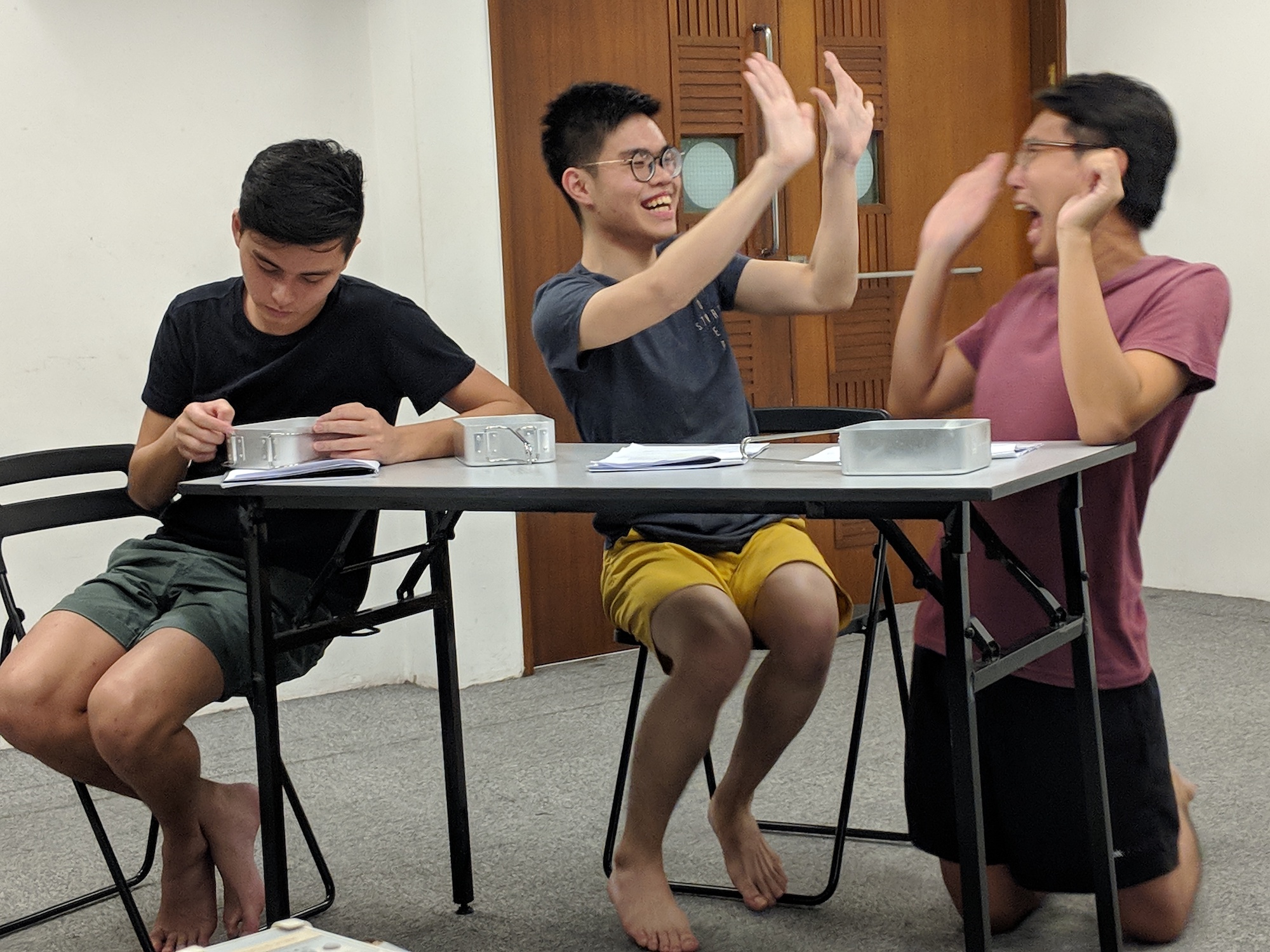 Three persons at a table, two excitedly high-fiving each other while the third looks pensively at a metal box in front of him.