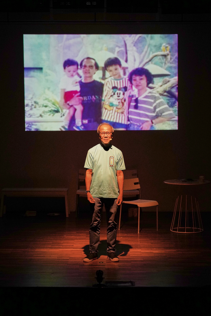 An elderly, male-presenting person standing under a spotlight. Behind him is an old-school family photograph projected on the wall.