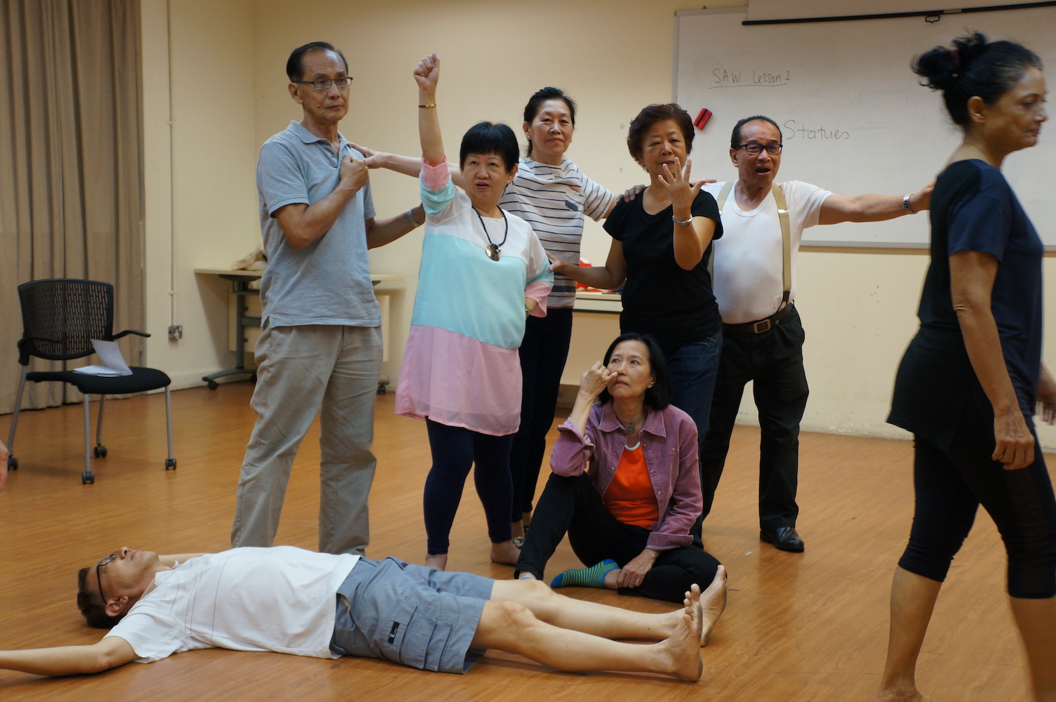 A group of six persons gesturing with their hands. In front of them is a male-presenting person lying face-up on the floor.