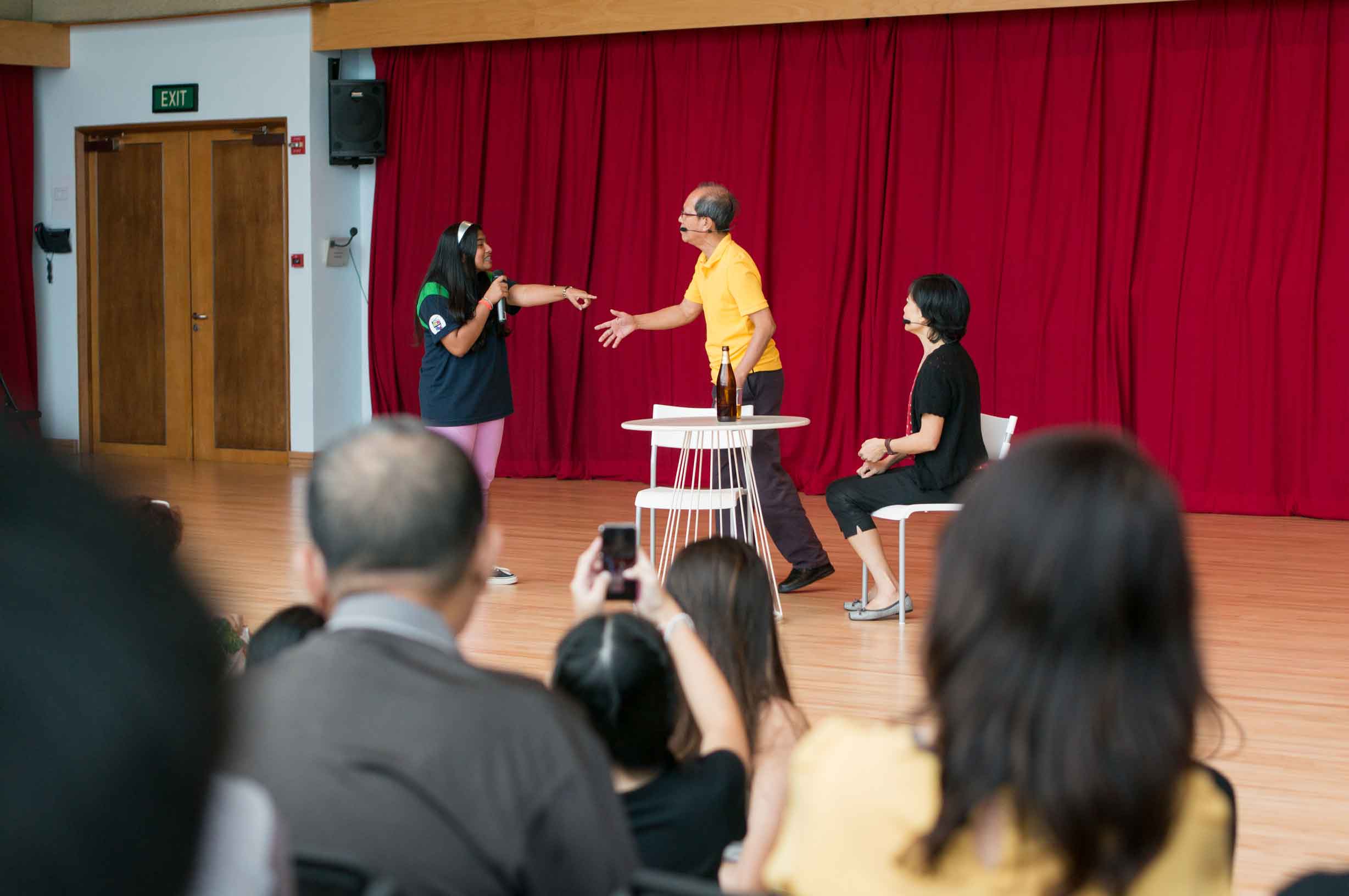 Three actors in front of an audience, two speaking and gesturing to each other.