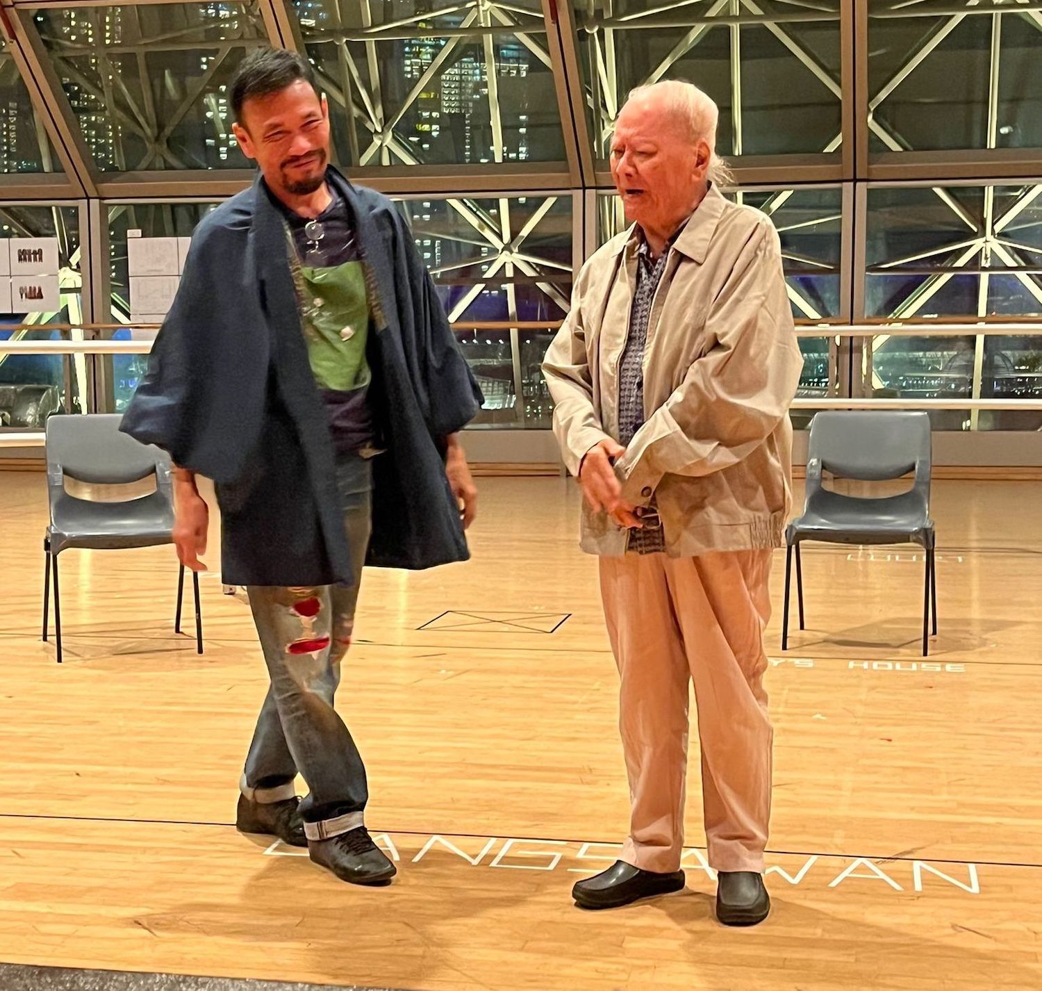 Two persons in jackets standing side by side in a room with a wood floor.