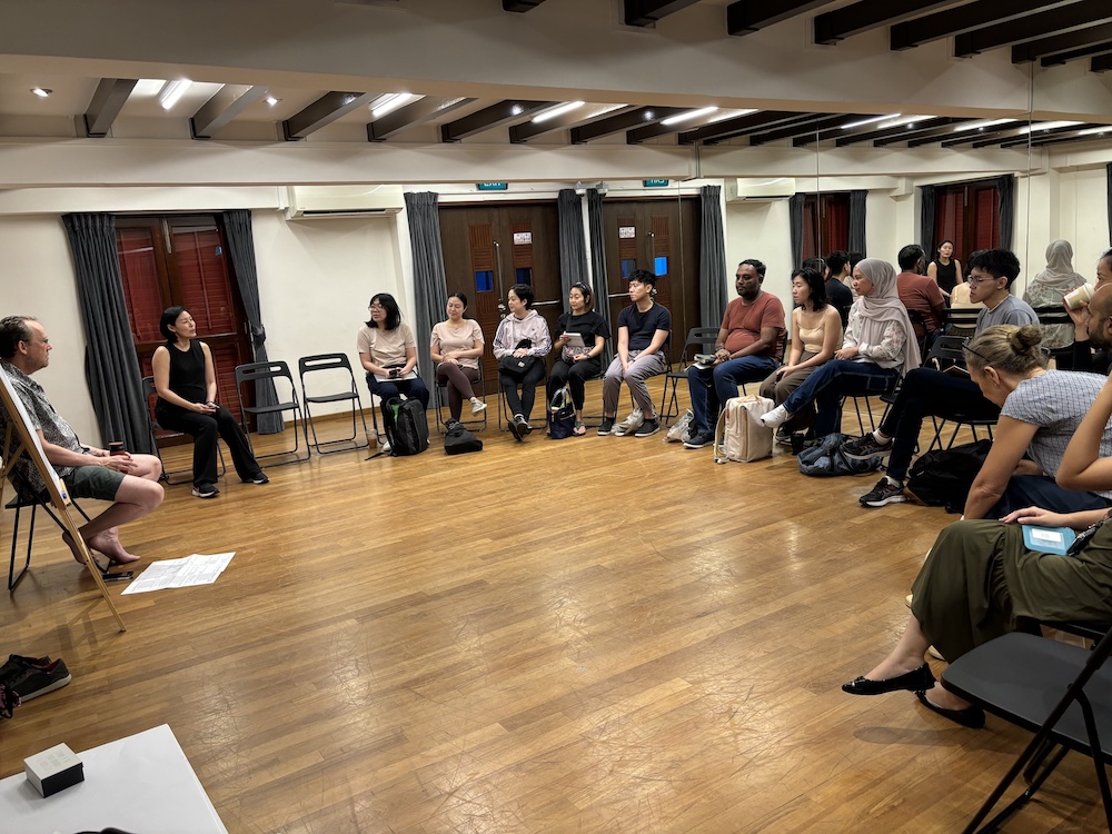 A semicircle of people seated in chairs, facing a person seated beside an easel.