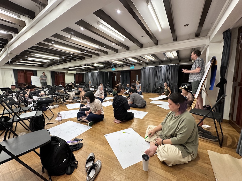 A room full of people seated on the floor with large sheets of paper, drawing and writing.