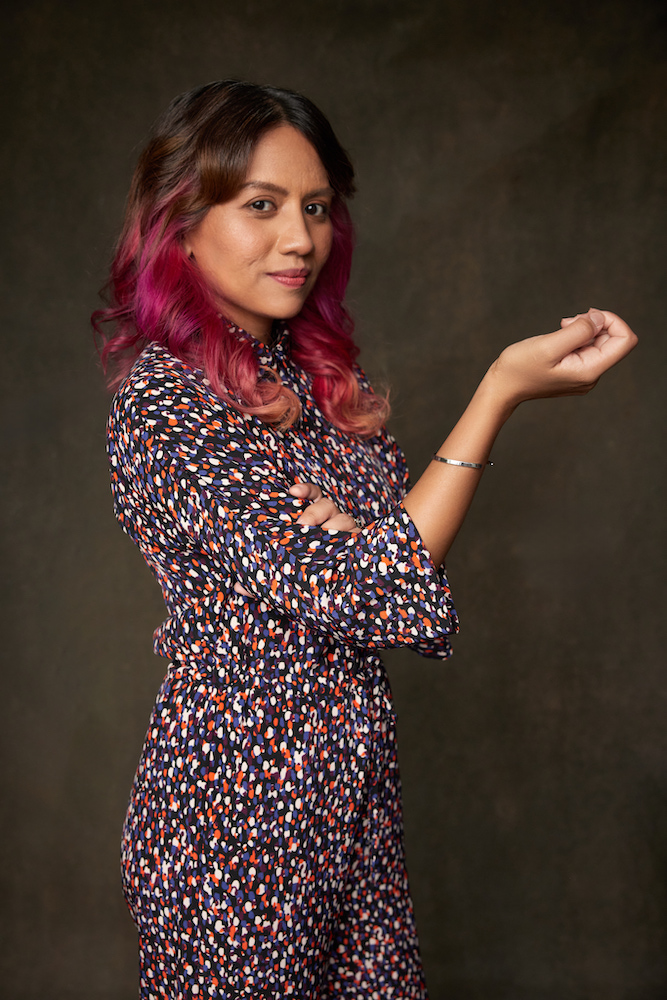 A female-presenting person in a colourful dress, right hand raised with her fist lightly clenched. A female-presenting person in a colourful dress, right hand raised with her fist lightly clenched.