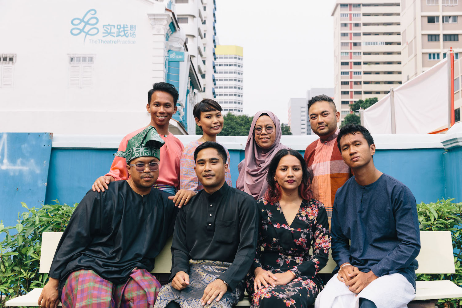 Eight people in bright, colourful outfits seated on a bench outdoors, smiling. Eight people in bright, colourful outfits seated on a bench outdoors, smiling.
