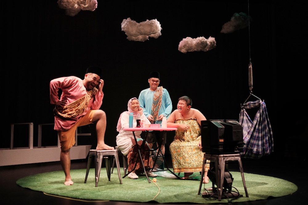 Four persons in colourful costumes in a spotlit area on a stage. Three are seated around a table smiling up at the last, who has his foot on a chair. Four persons in colourful costumes in a spotlit area on a stage. Three are seated around a table smiling up at the last, who has his foot on a chair.