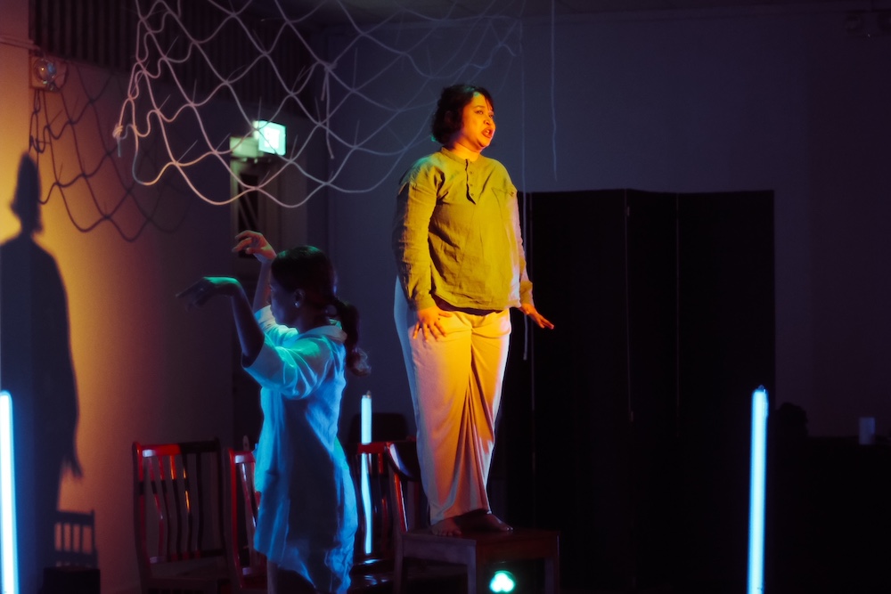 Two female-presenting persons - one standing on a chair, spotlit in yellow, and the other dancing to her left, spotlit in blue. Two female-presenting persons - one standing on a chair, spotlit in yellow, and the other dancing to her left, spotlit in blue.