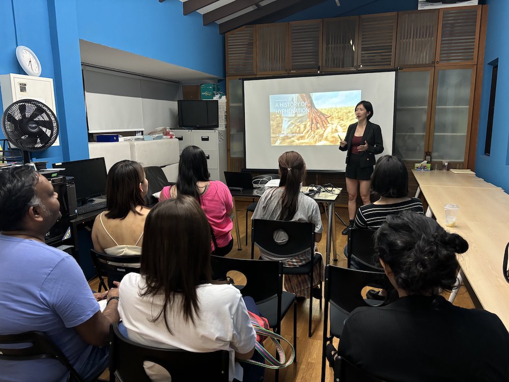 A photograph of the Centre 42 Office with attendees seated in rows of chairs, and Writer-in-Residence Jo Tan standing next to a screen with a slide that says "A History of Hyphenation".