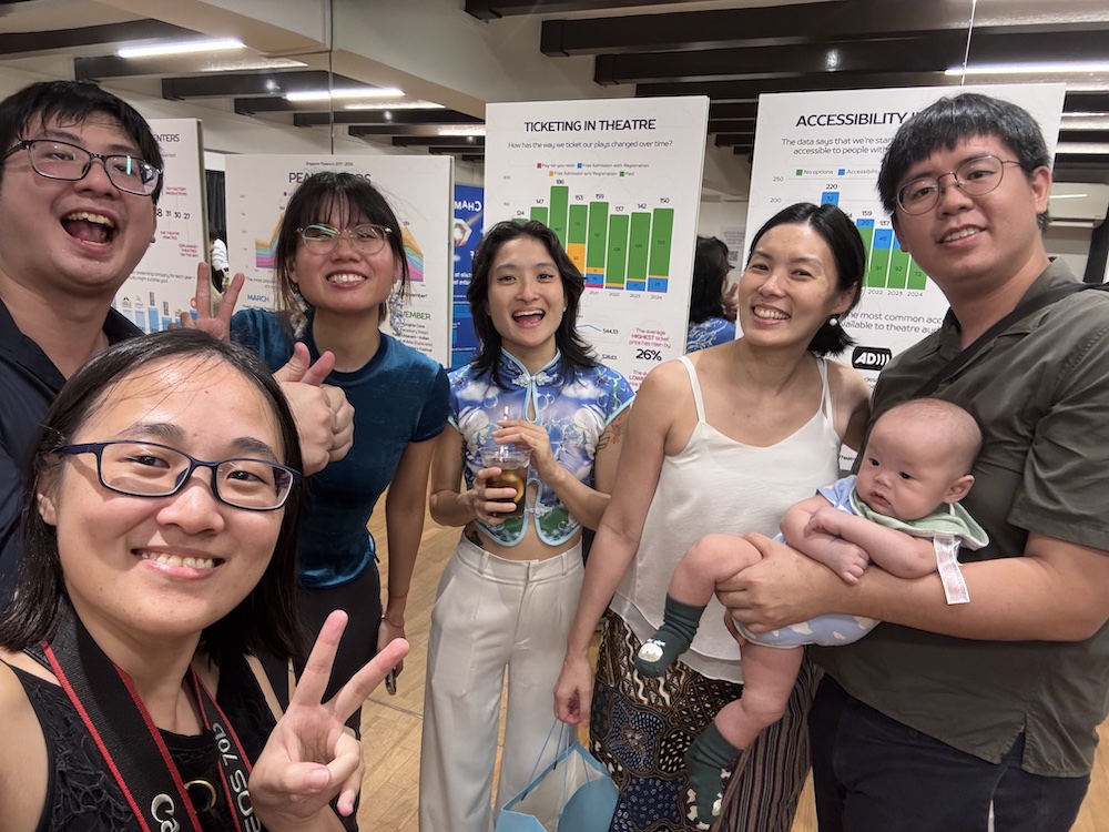 Six persons standing together and smiling into a camera. The rightmost person holds a baby in their arms.