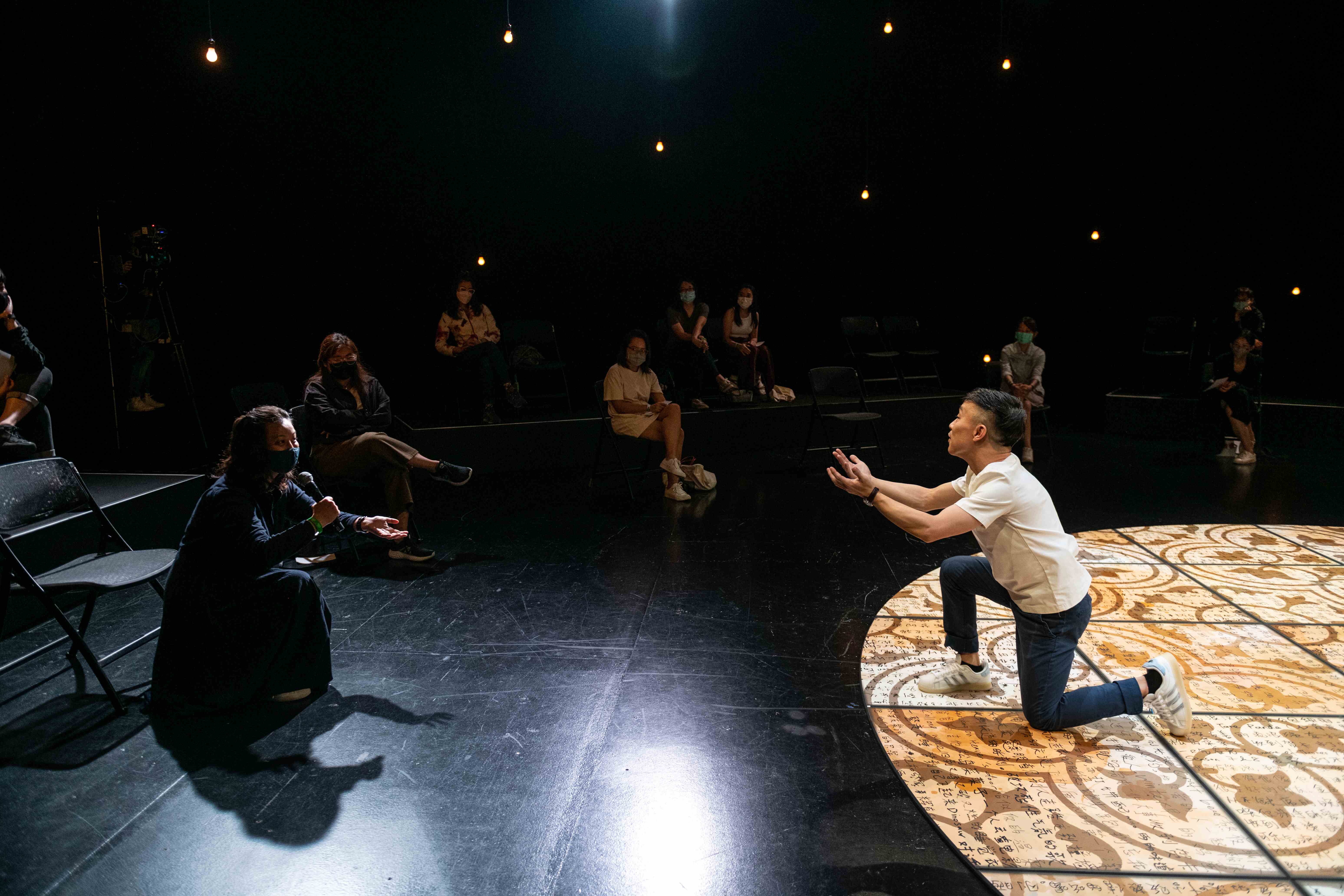 Performer, Oliver Chong, kneels on a patterned stage, reaching out with both hands in a dramatic gesture. An audience member is seen holding a microphone, watches and engages.