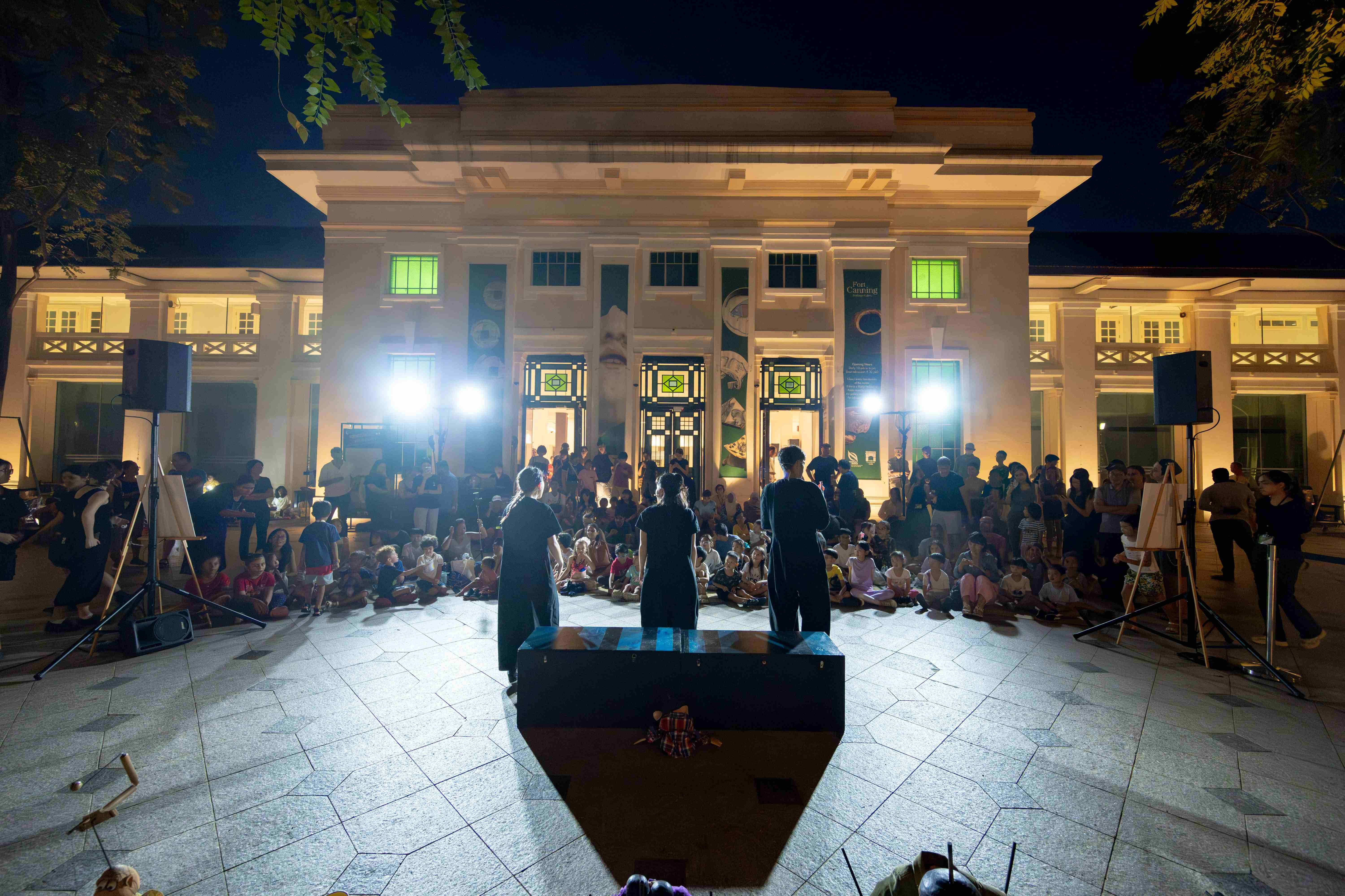 Three performers in black stand in front of a small black platform, facing a seated audience. The facade of Fort Canning Centre can be seen behind the audience.