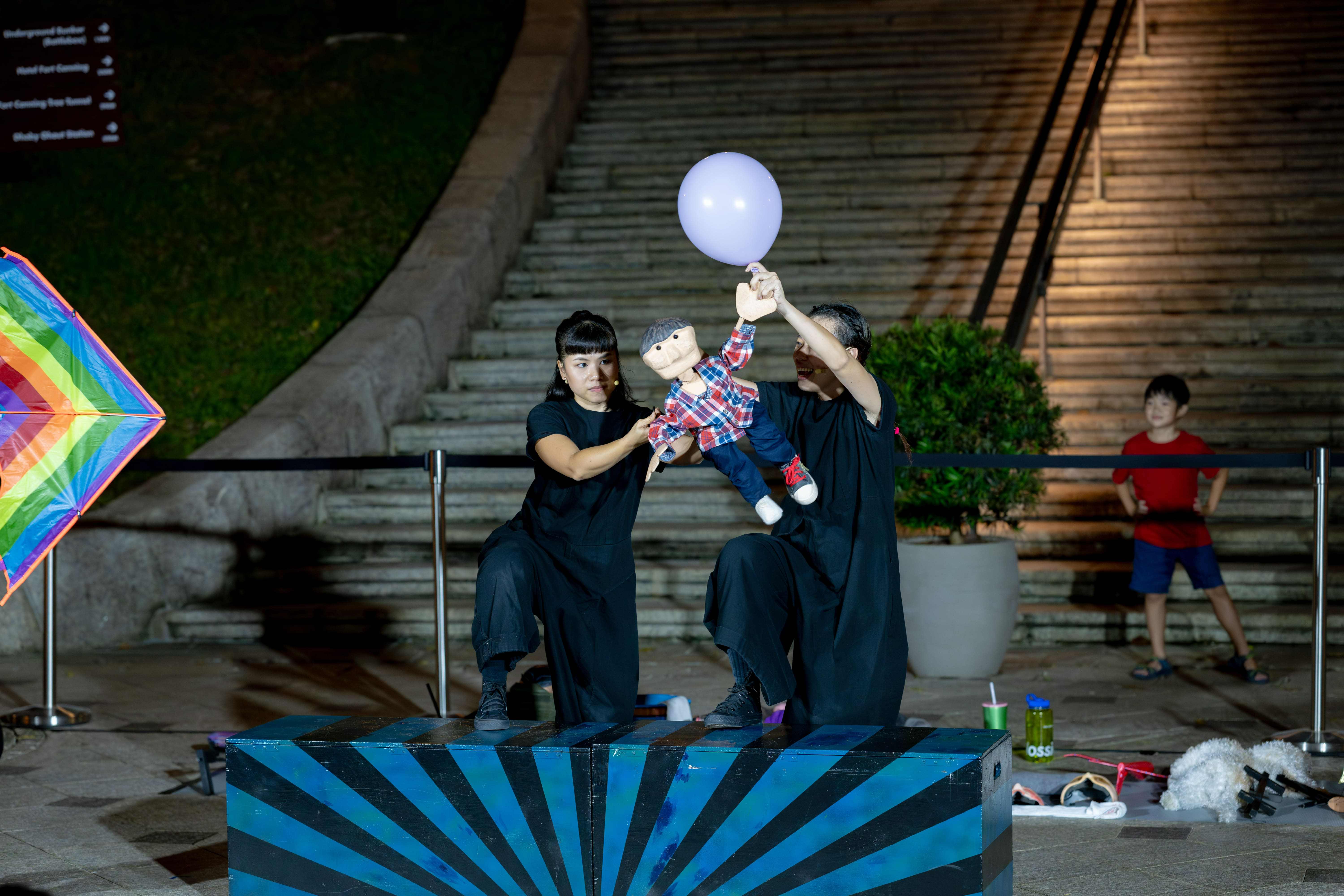 Two performers dressed in black manipulate a puppet holding onto a balloon on an outdoor stage.