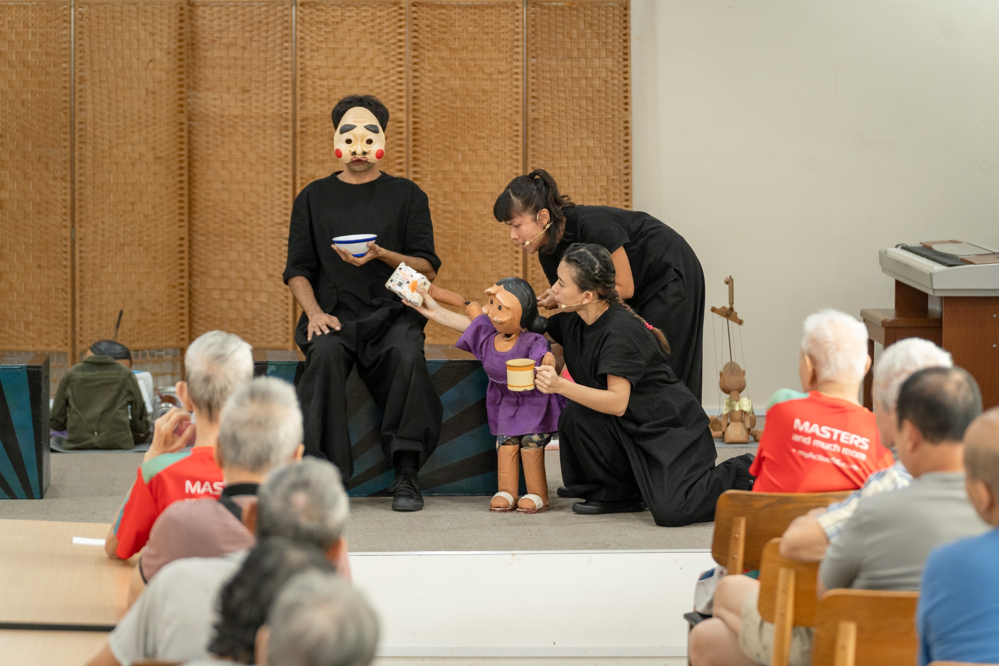 A masked puppeteer holds a bowl while sitting on a bench, as two puppeteers manipulate a small puppet, who points a tissue packet at the masked puppeteer while holding a cup. A number of elderly audience members can be seen in the foreground, out of focus.