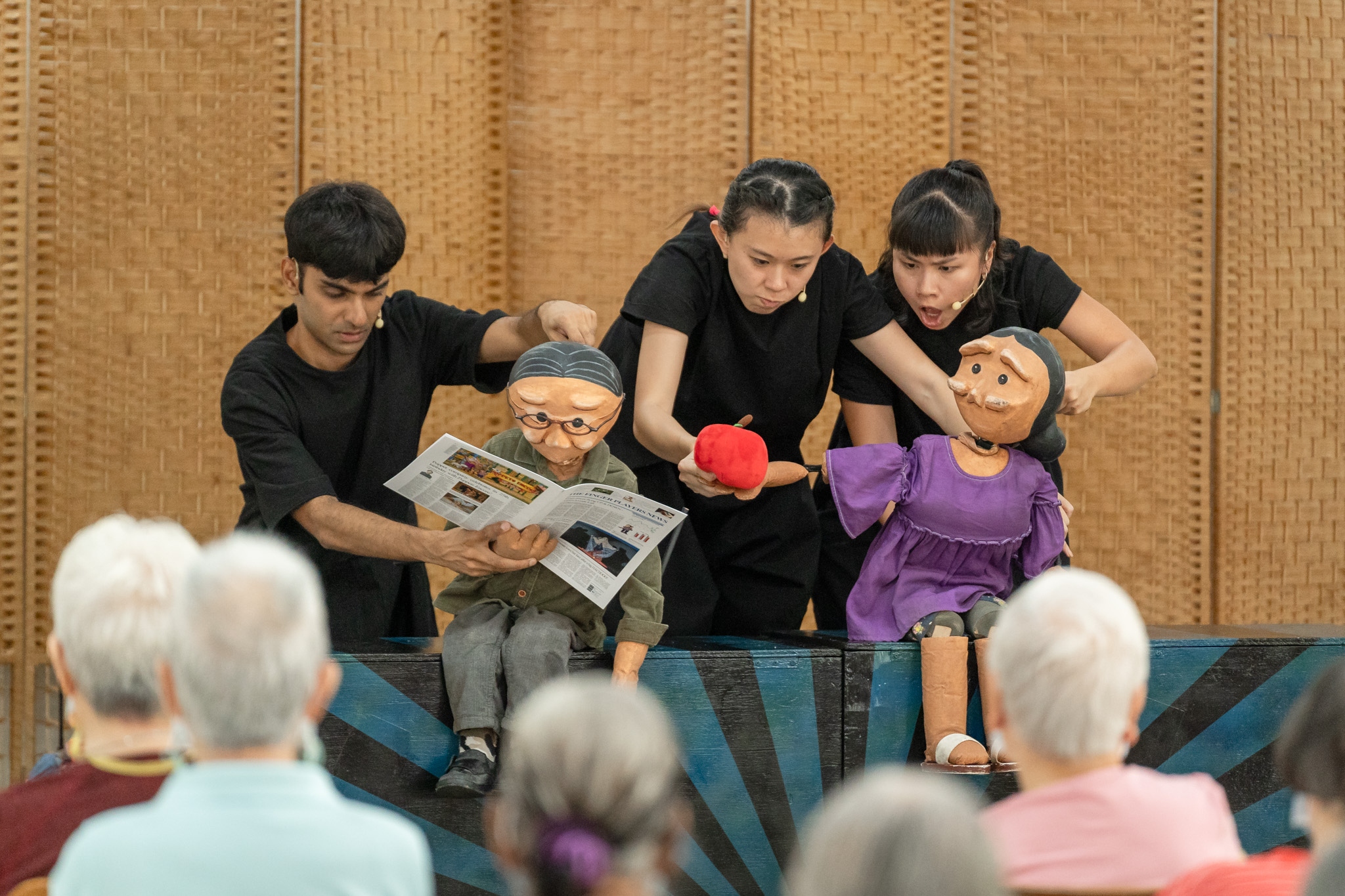 A puppeteer manipulates a small old man puppet which is reading a newspaper, while two puppeteers manipulate a small old woman puppet, who is handing the old man puppet an apple. A number of elderly audience members can be seen in the foreground, out of focus.