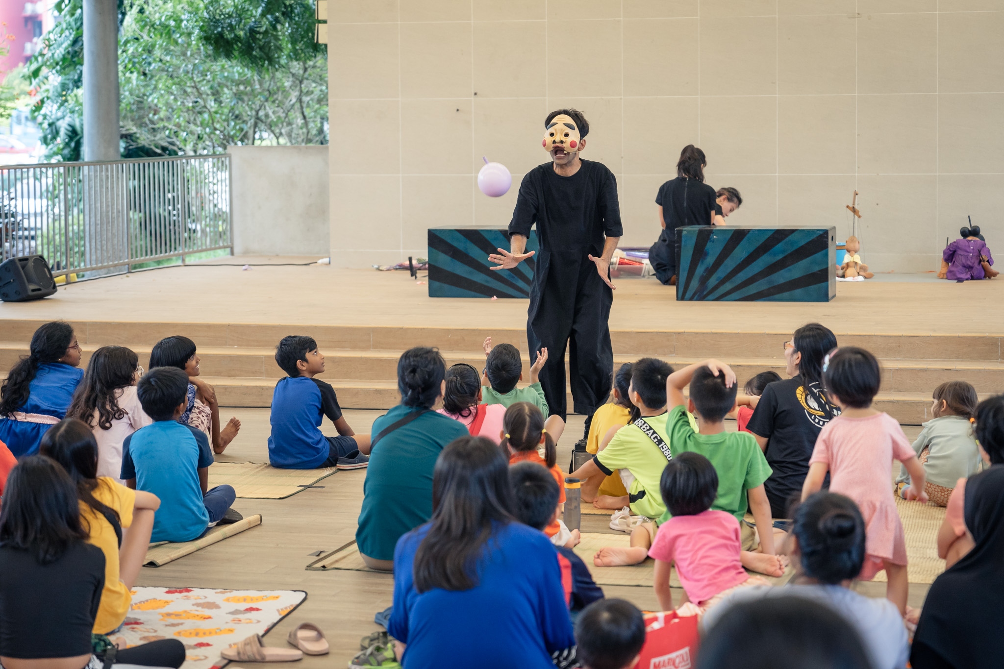 A masked puppeteer dressed in black clothes releases a balloon into the air as the seated audience composed mostly of children watches.
