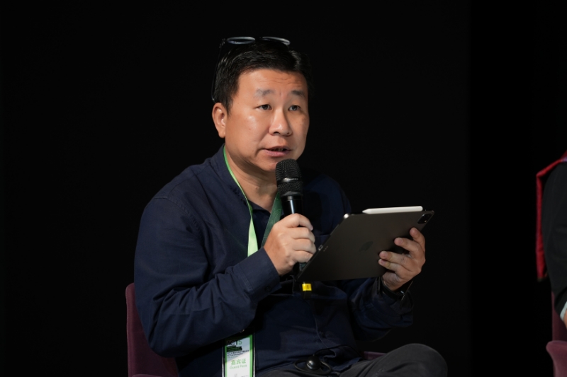 A male-presenting person speaking into a wireless microphone while holding a tablet. 