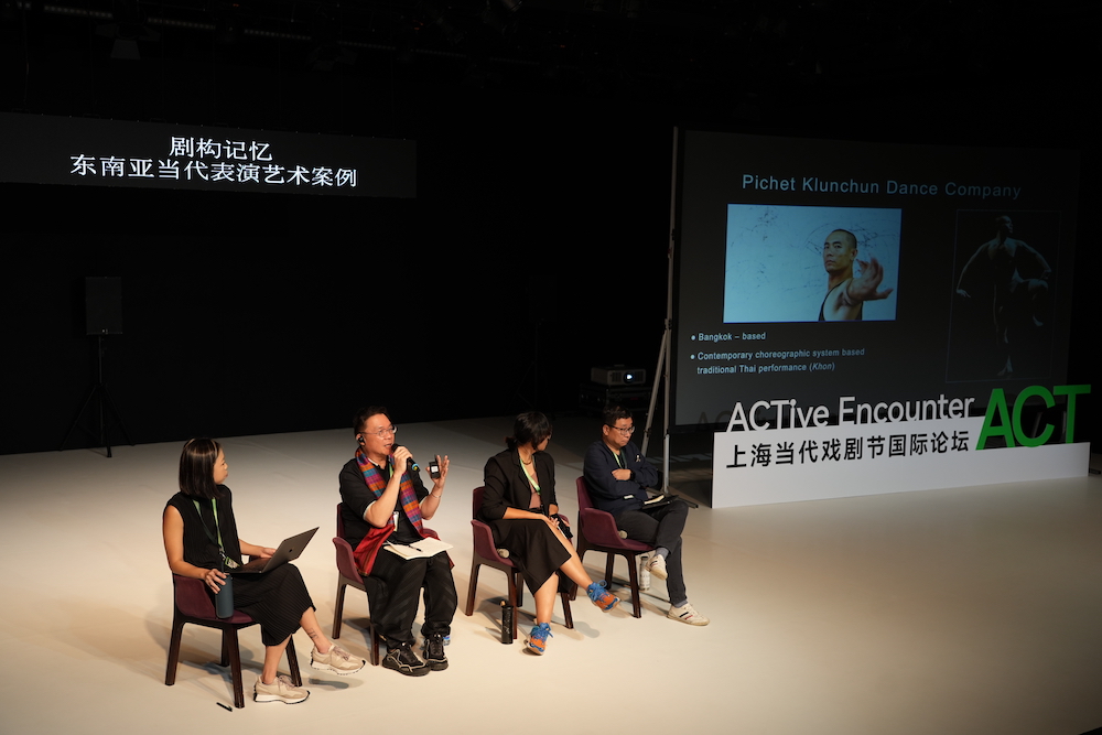 Four persons seated on a large white stage. The second person speaks into a microphone.