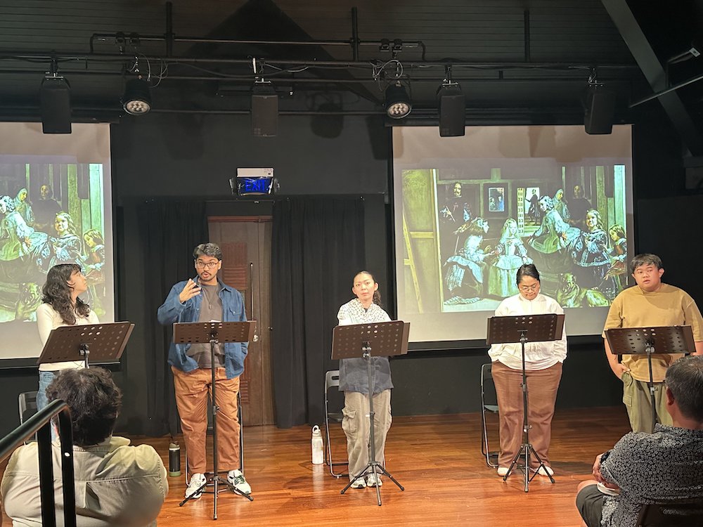 Five persons standing in a row in front of music stands. Behind them is a painting projected onto screens.