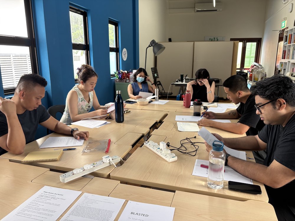 A group of persons seated around tables, reading from scripts.