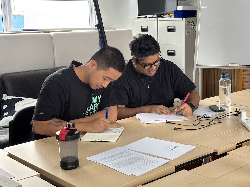 Two male-presenting persons bent over their notebooks and writing.