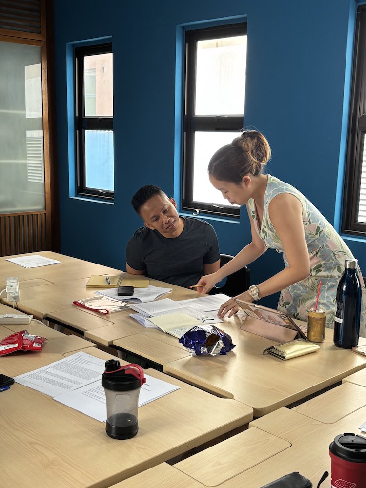 A female-presenting person stands, leaning over several papers. A male-presenting person is seated beside her, also looking at the papers.