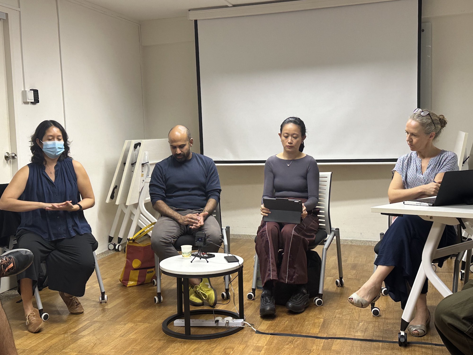 Four persons seated on chairs in a small room, in front of a small round white table. Four persons seated on chairs in a small room, in front of a small round white table.