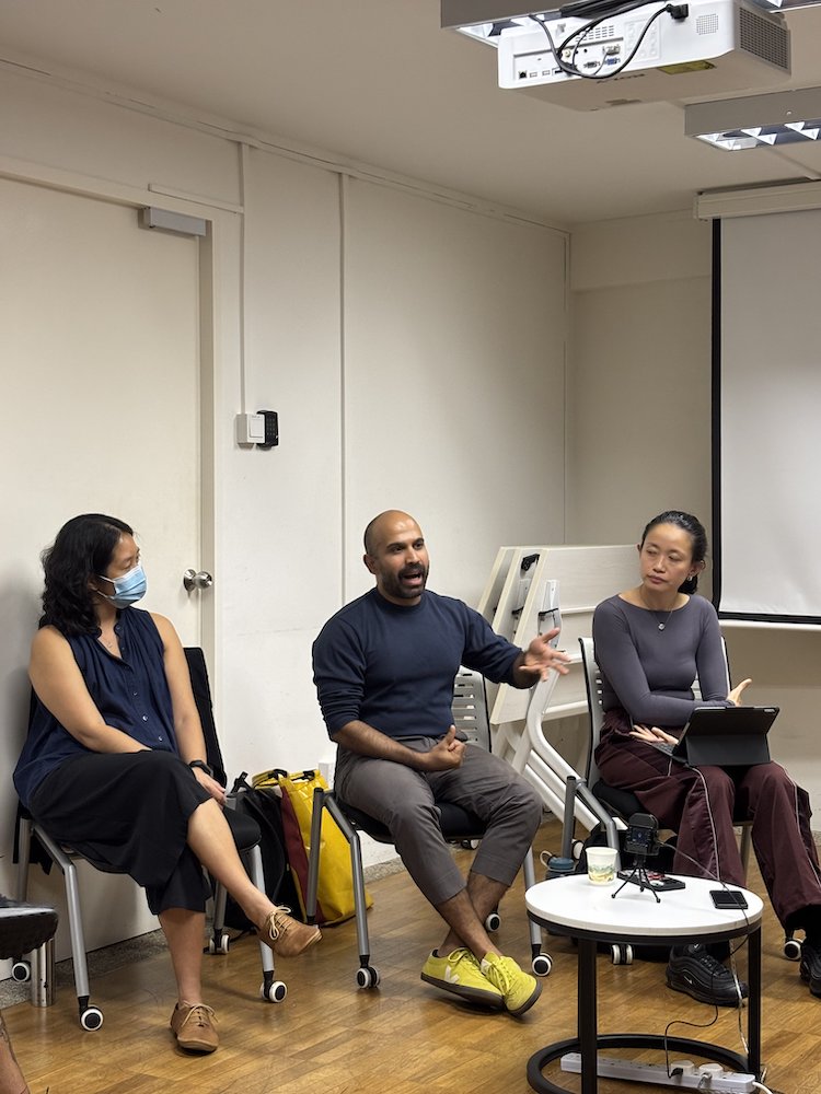 A male-presenting person seated between two female-presenting persons, speaking and gesturing. A male-presenting person seated between two female-presenting persons, speaking and gesturing.