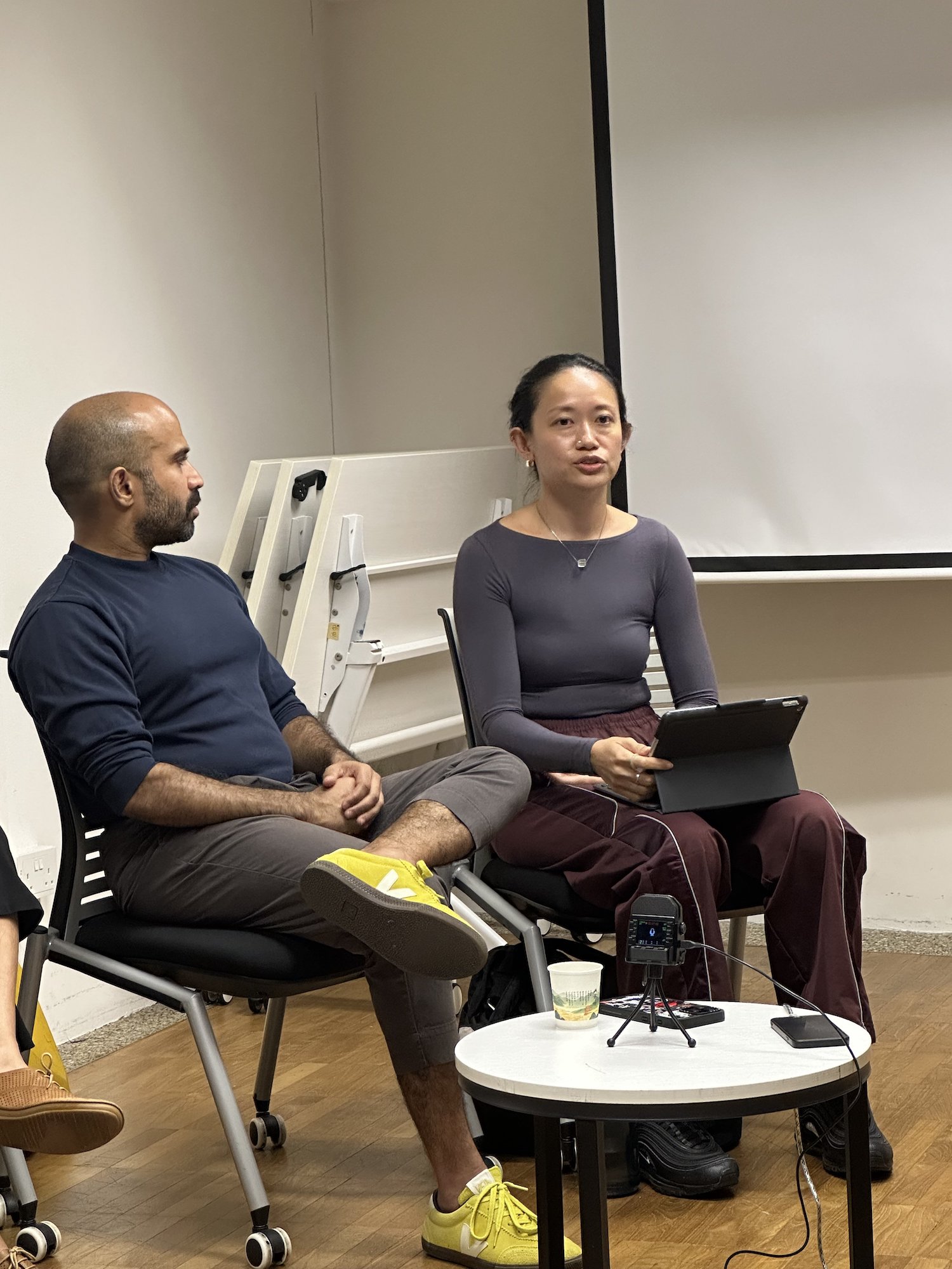 A female-presenting person seated on a chair, holding a tablet in her hands, and speaking. A female-presenting person seated on a chair, holding a tablet in her hands, and speaking.