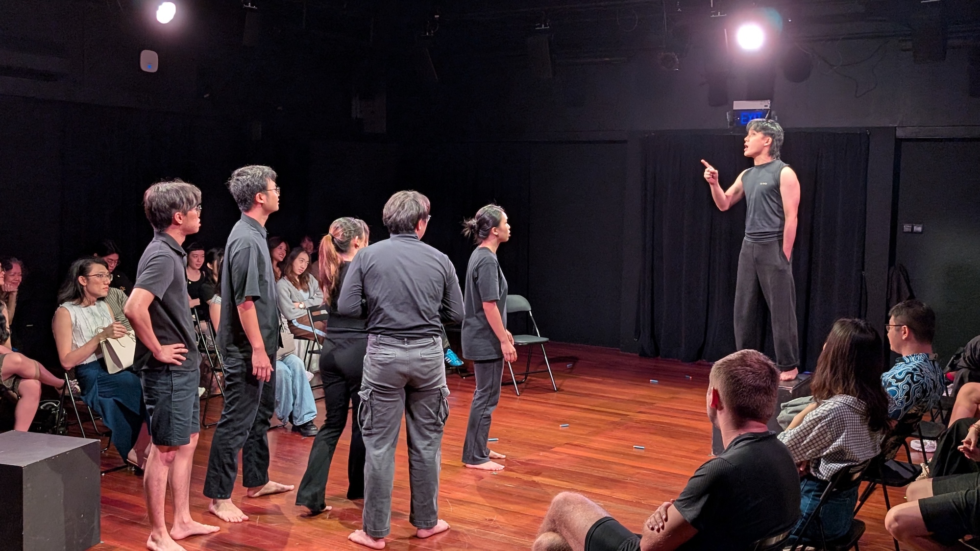 A male-presenting person standing on a box and pointing. Five persons in front of him stare up at him.