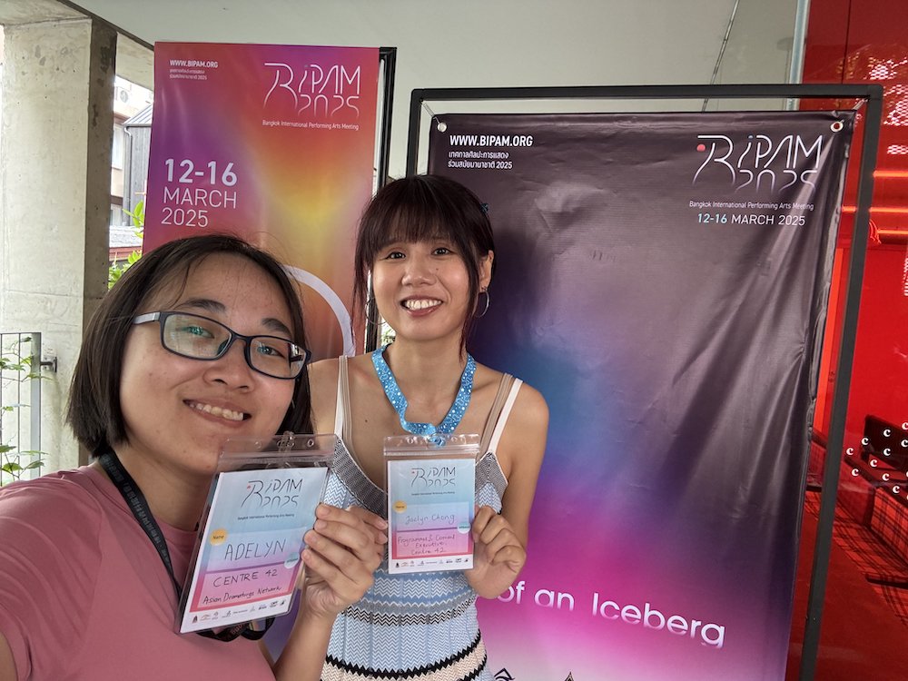 Two female-presenting persons standing in front of a purple banner, smiling and holding up two passes. Two female-presenting persons standing in front of a purple banner, smiling and holding up two passes.