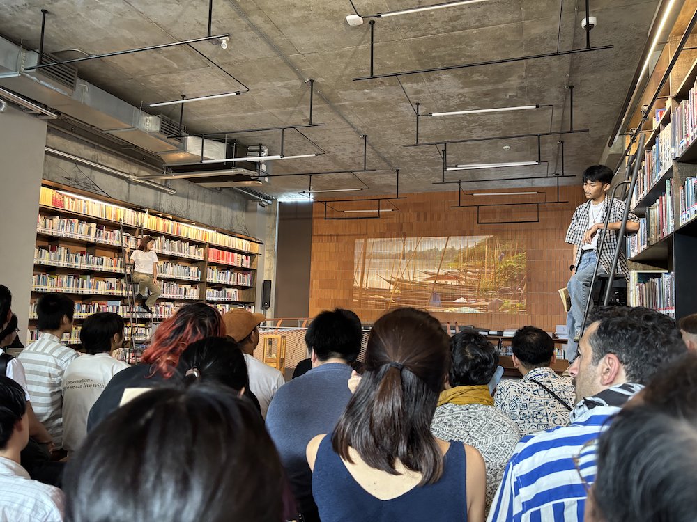 An audience watching two performers perched on top of ladders, leaned against full bookshelves. In the background an image is projected onto a brown wall. An audience watching two performers perched on top of ladders, leaned against full bookshelves. In the background an image is projected onto a brown wall.
