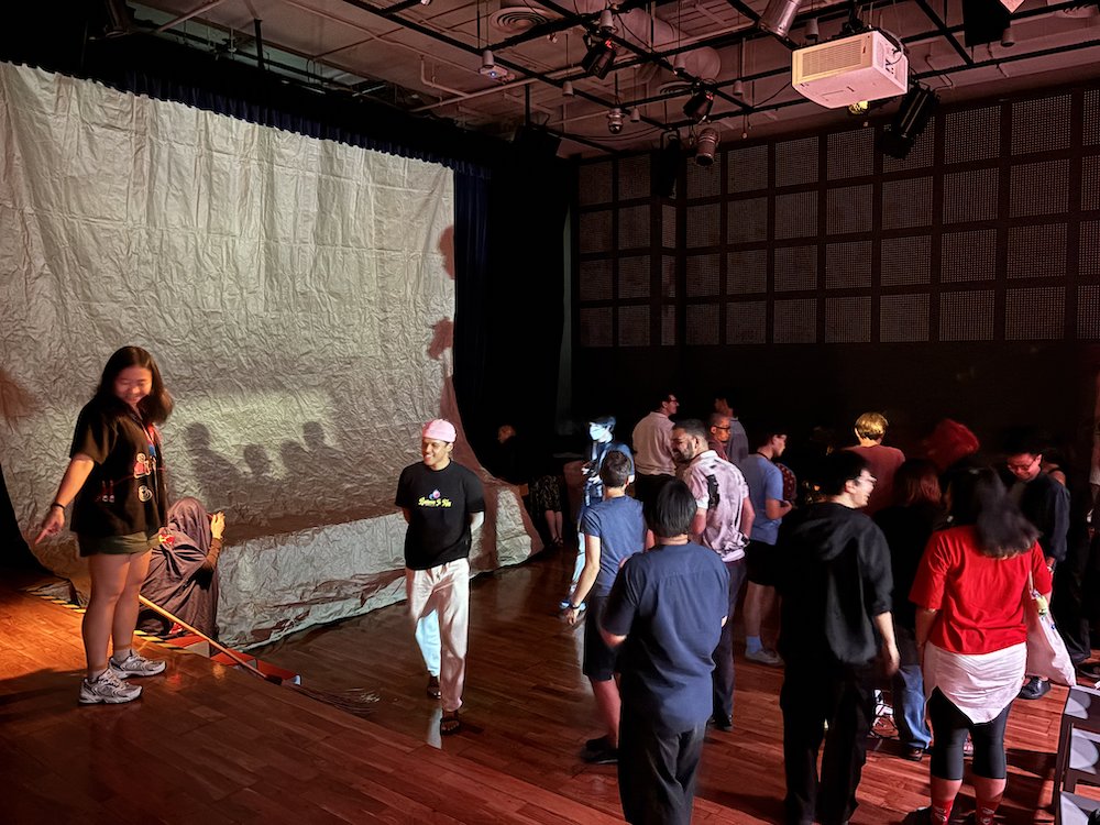 A large group of people milling around a dimly-lit theatre space. A large group of people milling around a dimly-lit theatre space.