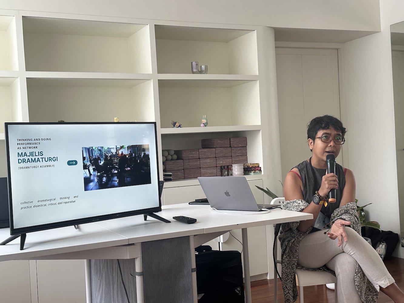 A female-presenting person speaking into a microphone. On the table beside her is a TV screen. A female-presenting person speaking into a microphone. On the table beside her is a TV screen.